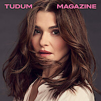 Rachel Weisz stands in front of a grey background wearing a white blouse, caught mid head turn with a clump of hair wrapping around her chin while looking to camera.
