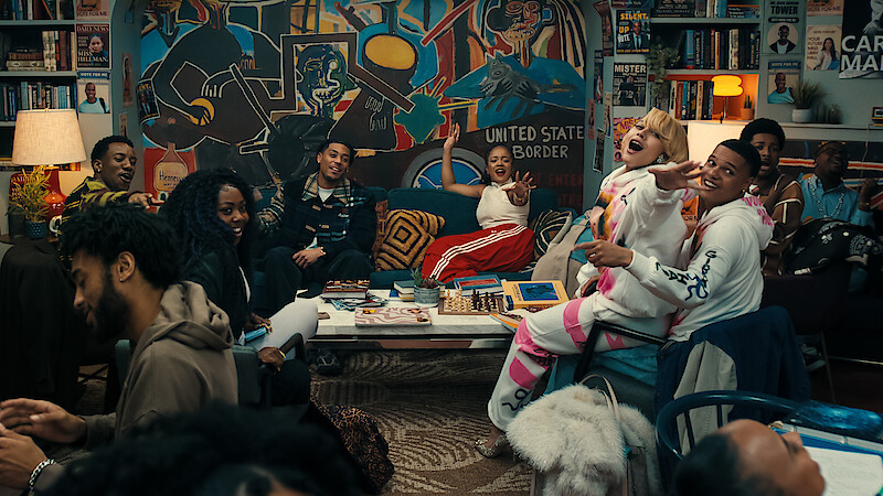 A diverse group of people laugh and socialize in a cozy, colorful living room filled with art, books, and a large mural, creating a lively, welcoming atmosphere.