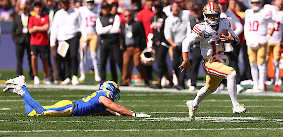 Football quarterback in a white and gold uniform runs with the ball on a sunny field while a defender in a blue and yellow uniform dives to tackle him; crowd and teammates watch in the background.