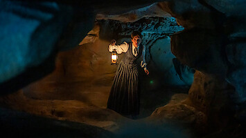 Woman in historical clothing holding a lantern exploring a dimly lit rocky cave, surrounded by shadows and uneven cave walls, with a mysterious blue light in the background.