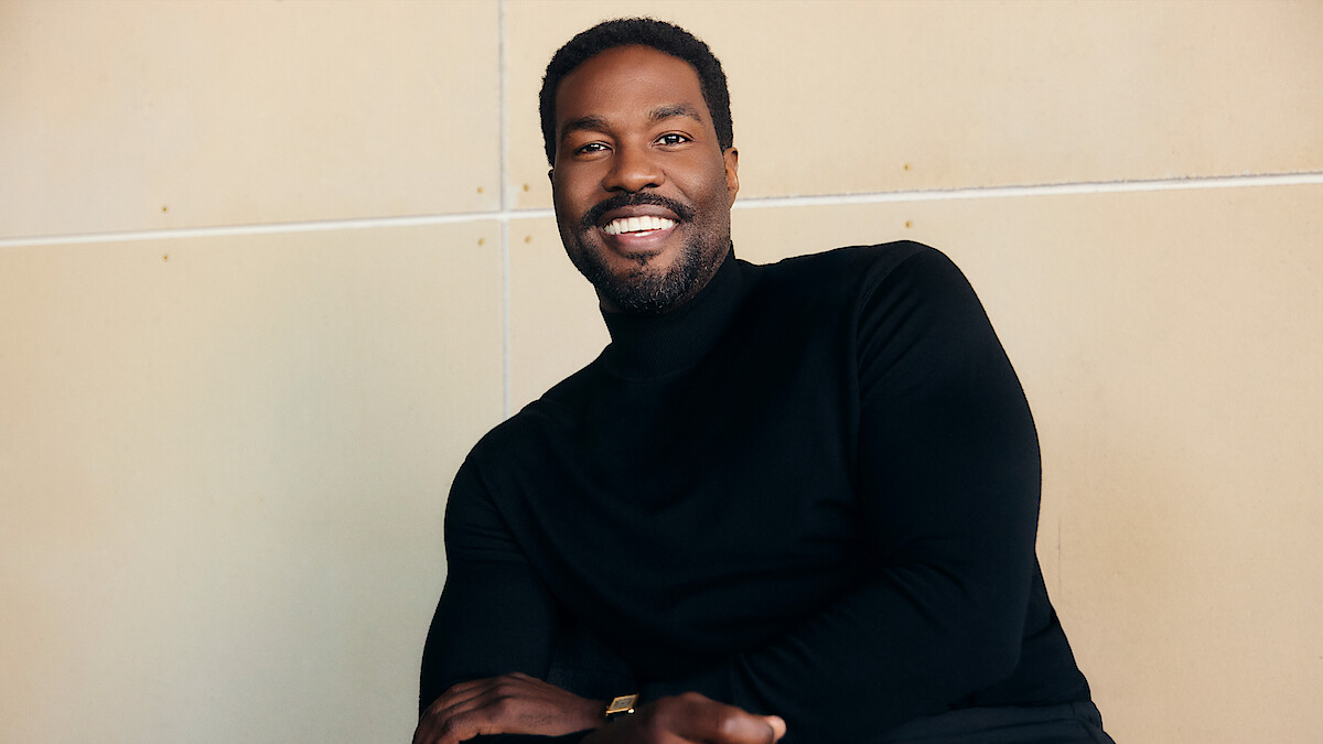 Man in black turtleneck sweater sitting and smiling in front of a light-colored wall in a casual indoor setting.
