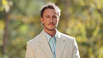 Drew Starkey em pé usando camisa azul e blazer claro com textura em um ambiente externo ensolarado, diante de um plano de fundo esverdeado desfocado que sugere um ambiente natural e tranquilo.