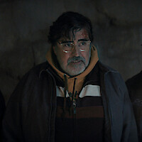 An older man with glasses and a beard, wearing a hoodie and jacket, stands indoors against a dimly lit, textured wall, looking slightly to the side.
