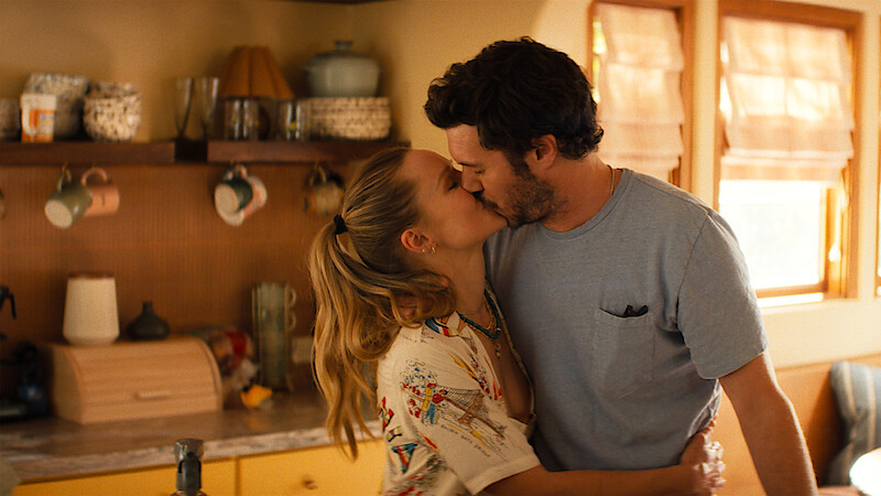 A couple kisses in a warm, sunlit kitchen with wooden cabinets, shelves holding mugs and jars, and soft natural light coming through the window.