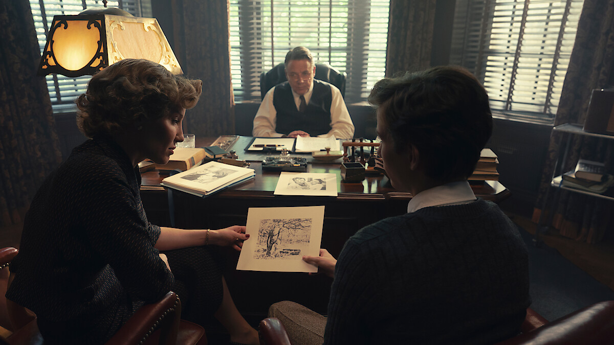 Two people sit across from a man at a desk in a dimly lit office, discussing a drawing. The office has large windows with blinds, books, and papers scattered on the desk, creating a serious, professional atmosphere.