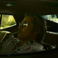 Man wearing sunglasses sits in the driver’s seat of a car, viewed from the side window, with sunlight filtering in. The car interior has a retro style and relaxed vibe.