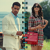 A man in a white suit and a woman in a tweed dress with a red handbag stand outdoors, split by a vertical blue graphic, with a lake and greenery in the background. Both appear stylish and fashionable.