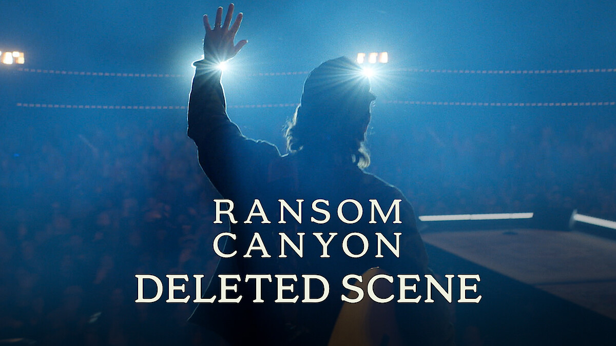 Thomas Rhett stands on stage in silhouette, facing bright spotlights with one hand raised. Smoky blue lighting fills the scene, the audience is barely visible in the background, and a text overlay reads: "Ransom Canyon Deleted Scene."