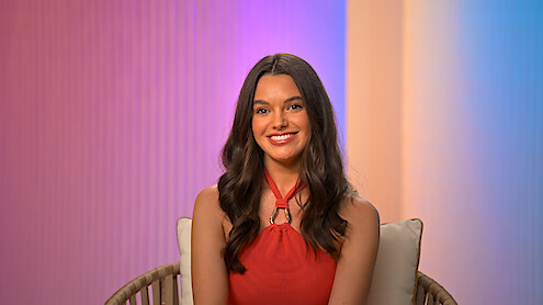 Ally with long brown hair in a red halter dress sitting on a chair, smiling in a studio with pastel gradient background lighting.