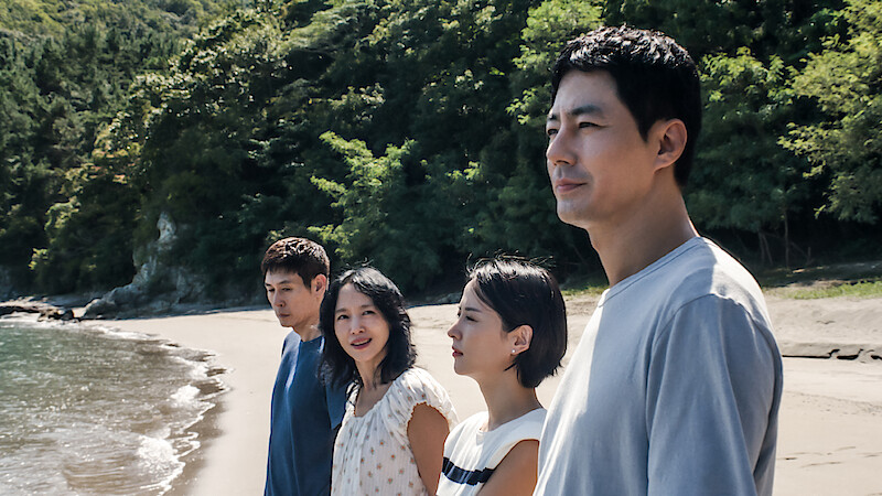 Four people stand side by side on a sandy beach with trees and greenery in the background, looking thoughtful under natural daylight by the shoreline.