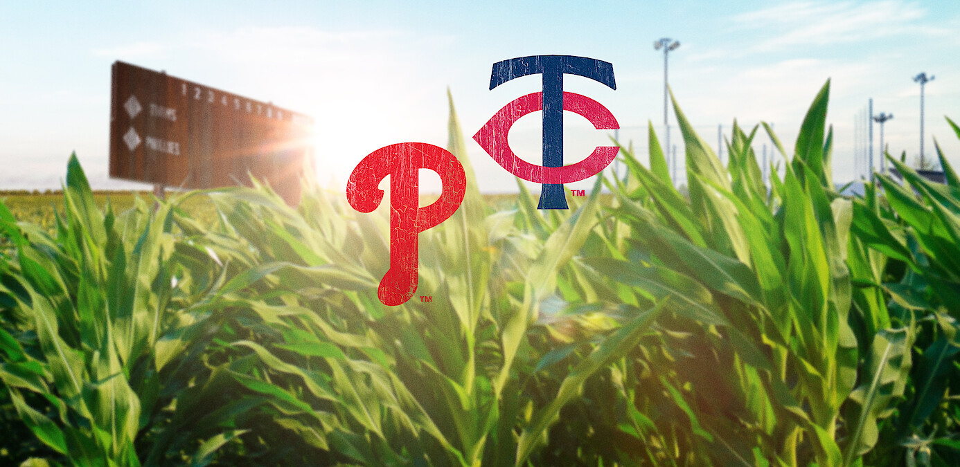 Baseball field scoreboard and stadium lights in the background, lush green cornfield in foreground, with Philadelphia Phillies and Minnesota Twins logos superimposed in the center, sunlight shining through.