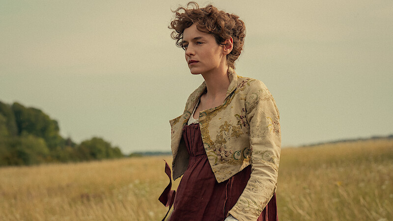 Woman in historical dress standing in a field with tall grass, trees in the background, and a cloudy sky, creating a calm, pastoral setting.