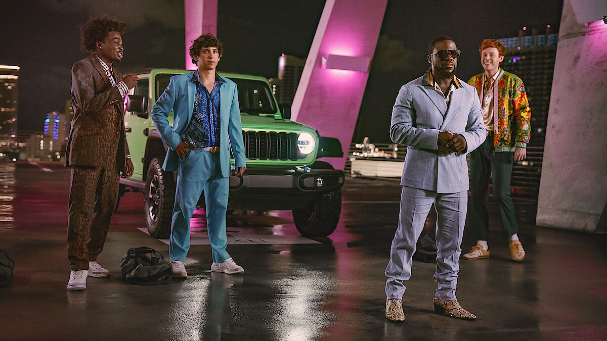 Four men in stylish suits stand in front of a green Jeep on a rooftop at night, illuminated by pink and purple lights, with a cityscape in the background.