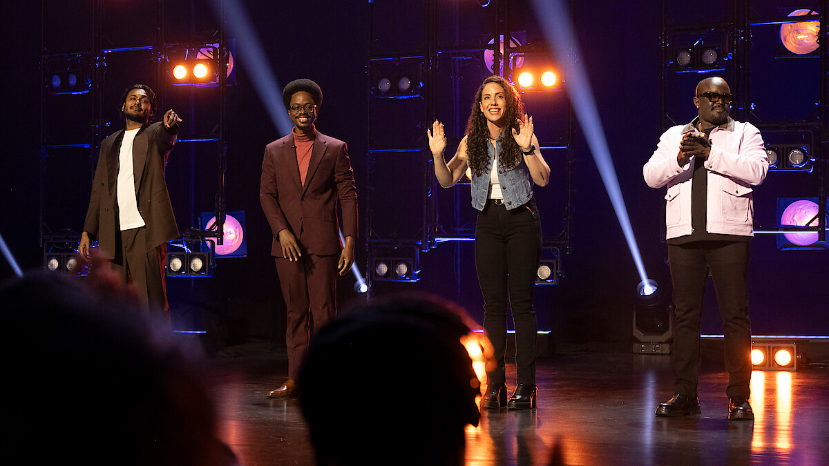 Four people stand on a stage with bright lights and spotlights behind them, in a lively performance or show setting, with one person pointing, two others smiling, and the group facing an audience.