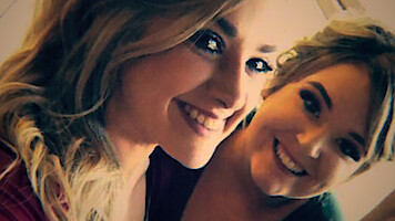 Two young women smiling closely together, looking at the camera indoors with soft, warm lighting. Casual atmosphere, both appear happy and relaxed.