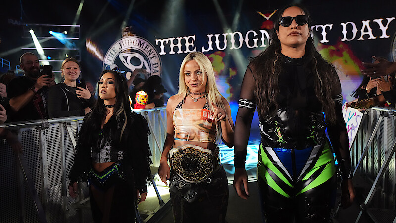 Three female wrestlers walk down an arena entrance ramp surrounded by cheering fans, with one in the center holding a championship belt. Bright lights and "The Judgment Day" appear in the background.