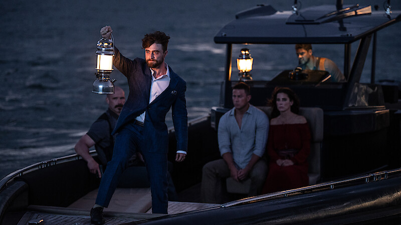 A man in a blue suit holds up a lantern on a small boat at dusk, surrounded by four other people sitting and watching, with a dark sea and another boat in the background.
