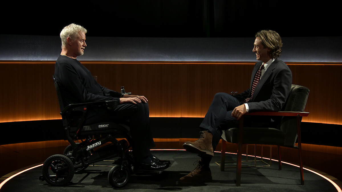 Eric Dane, em uma cadeira de rodas, e Brad Falchuk, em uma cadeira, estão frente a frente em um estúdio com iluminação suave. Eles conversam em um palco com um fundo minimalista.