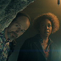 Two people in dim lighting, one peering around a rocky edge and the other standing, both looking intently ahead. The environment suggests a cave or underground setting with a single hanging lightbulb.