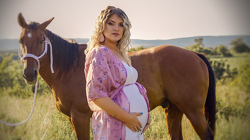 Pregnant woman in a pink floral robe stands in a grassy field, gently holding her belly, next to a brown horse with a white halter. Background shows rolling hills under a sunny sky.