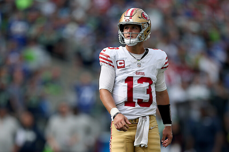 Football player wearing a San Francisco 49ers uniform and helmet stands on the field during a game, with a blurred crowd and stadium setting in the background.