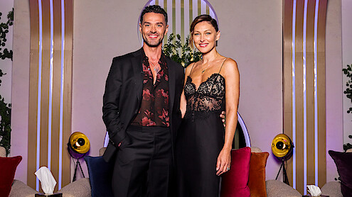 Homem de terno preto e mulher com vestido preto de renda estão juntos, sorrindo, em uma sala de estar moderna com almofadas coloridas, luminárias redondas, plantas e linhas verticais decorativas ao fundo.