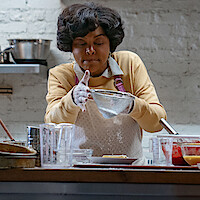 A woman in a kitchen sifts flour over a dish, surrounded by glassware and bowls on a wooden counter, with a white brick wall and metal shelf in the background, creating a cozy, homey cooking environment.