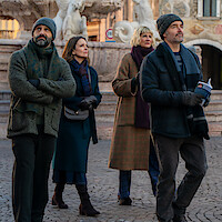Four adults in winter clothing walk together in a cobblestone plaza, with an ornate fountain and historic buildings in the background, suggesting a city sightseeing setting in cold weather.