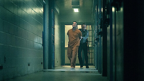 A man in an orange prison jumpsuit is being escorted by a guard down a dimly lit hallway in a jail or prison, with barred cells and concrete walls visible.