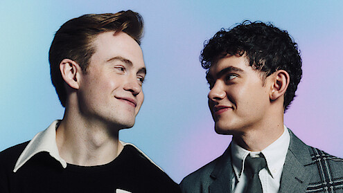 Two young men smiling warmly at each other, one in a casual shirt and the other in a suit and tie, with a soft blue and purple gradient background. Studio lighting, friendly atmosphere.