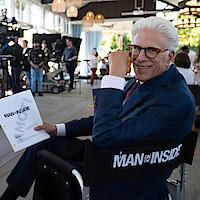 Smiling man in a suit sits on a director’s chair labeled "Man on the Inside," holding a script on a bright, busy film set with crew and equipment in the background.