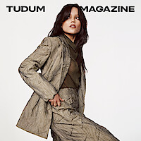 Jenna Ortega in a taupe textured suit with sheer blouse looks to the side, gaze locked on something in the distance with her hair down.