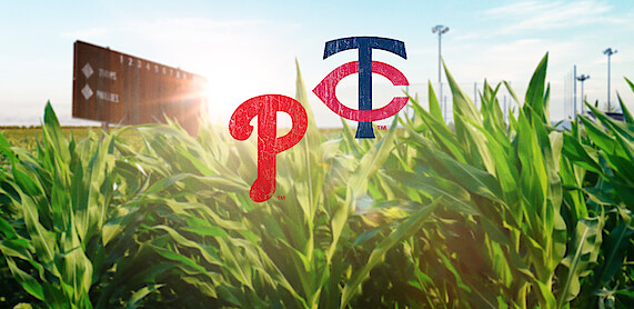 Baseball field scoreboard and stadium lights in the background, lush green cornfield in foreground, with Philadelphia Phillies and Minnesota Twins logos superimposed in the center, sunlight shining through.