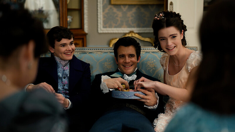 A joyful group of people in period clothing sit closely on a blue patterned couch in an ornate living room, smiling and sharing snacks, with antique furniture and elegant decor in the background.