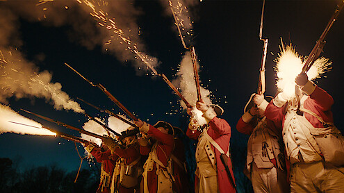 Historical reenactors in red coats fire muskets with bright flashes in a nighttime outdoor setting, capturing an intense moment in a dramatic historical battle scene.
