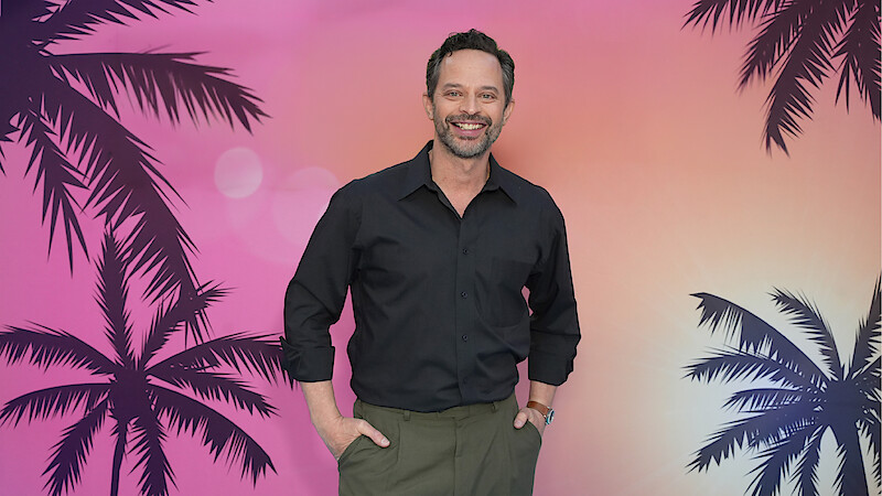 A smiling man in a black shirt and green pants stands in front of a colorful sunset backdrop with palm tree silhouettes, evoking a tropical or beach setting.