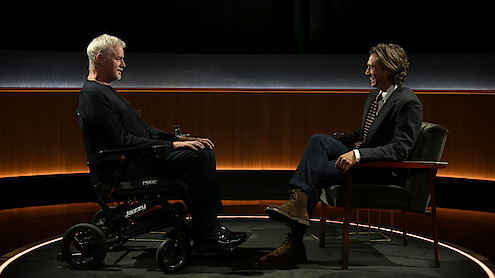 Eric Dane, em uma cadeira de rodas, e Brad Falchuk, em uma cadeira, estão frente a frente em um estúdio com iluminação suave. Eles conversam em um palco com um fundo minimalista.