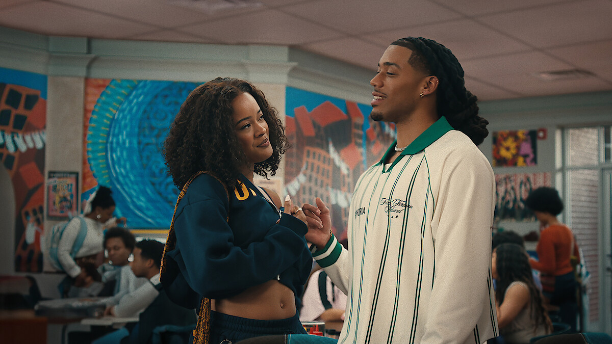 Two young adults talking closely in a colorful school cafeteria, with other students sitting and interacting in the background, murals and bright artwork on the walls.