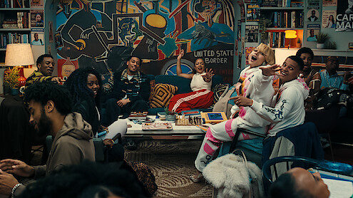 A diverse group of people laugh and socialize in a cozy, colorful living room filled with art, books, and a large mural, creating a lively, welcoming atmosphere.