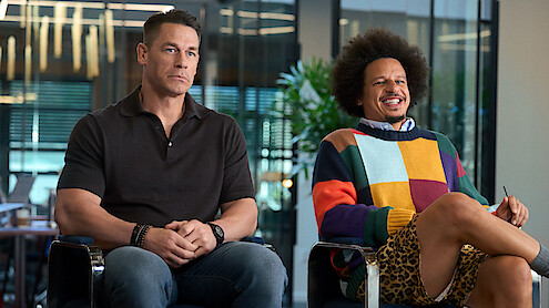 Two men sit indoors on chairs, one in a black polo shirt looking serious, the other in a colorful sweater and animal-print shorts smiling, with plants and modern office glass walls in the background; mood is contrasting and casual.