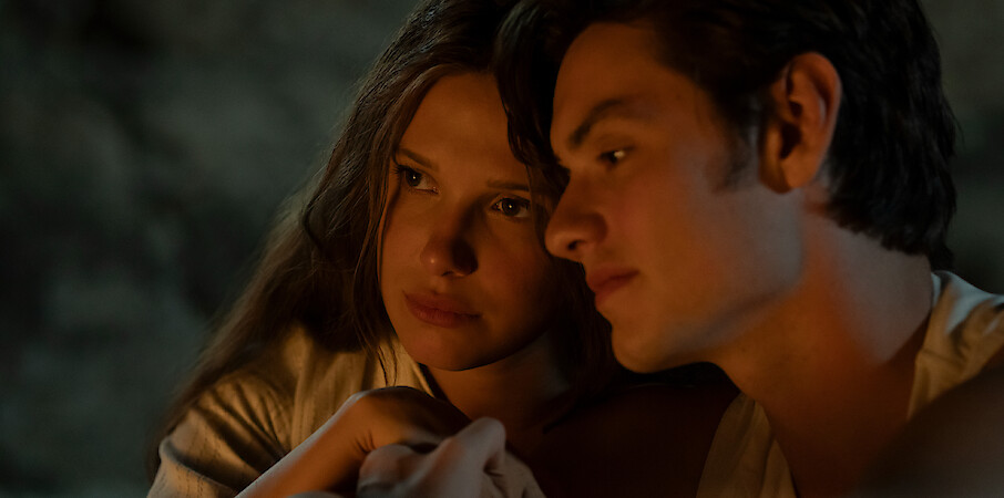 Two young people sit closely together in a dimly lit, cozy setting, with warm light illuminating their faces, suggesting an intimate or reflective moment.