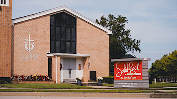 Igreja de tijolos com grande vitral preto, logotipo com cruz e coração e placa "Solid Rock" na fachada. Gramado bem cuidado, flores e árvores em um ambiente suburbano sob um céu claro.