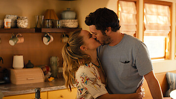 A couple kisses in a warm, sunlit kitchen with wooden cabinets, shelves holding mugs and jars, and soft natural light coming through the window.