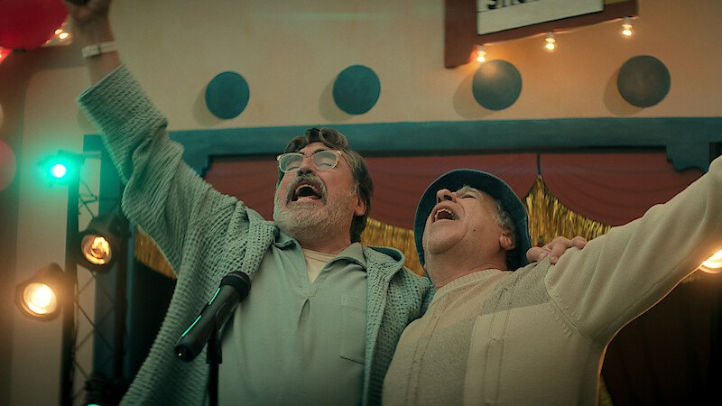 Two older men joyfully sing together on stage with arms raised, surrounded by stage lights and festive decorations in an indoor event setting, suggesting a celebratory or performance atmosphere.