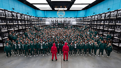 Hundreds of people in green tracksuits stand in rows inside a large, brightly lit room with bunk beds, facing two guards in pink uniforms.