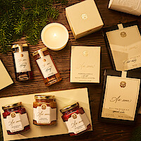 A cozy, warm-toned flat lay of assorted elegantly-packaged gift items, including candles, jars, boxes, and honey, arranged on a wooden table with pine branches, evoking a festive and inviting holiday mood.