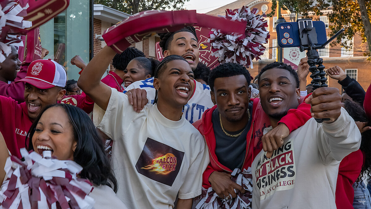 Group of college students smiling, cheering, and taking a selfie outdoors at a lively campus event, wearing school colors and holding pompoms, with historic campus buildings in the background.