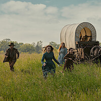 A family runs through a field with a wagon behind them. 