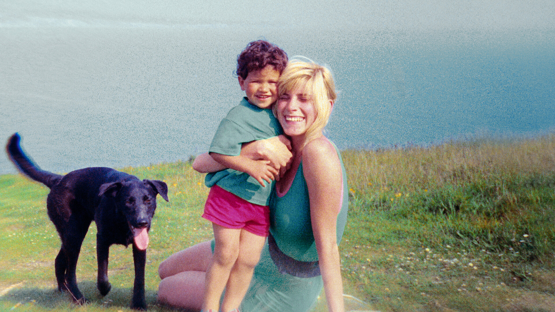 Smiling woman sits in grass near water, hugging a young child while a black dog stands nearby outdoors on a bright day.