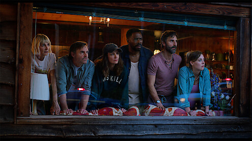 Six people lean forward together at a wooden cabin window, looking outside with concerned and curious expressions; the cozy, warmly lit interior contrasts with the dramatic mood.